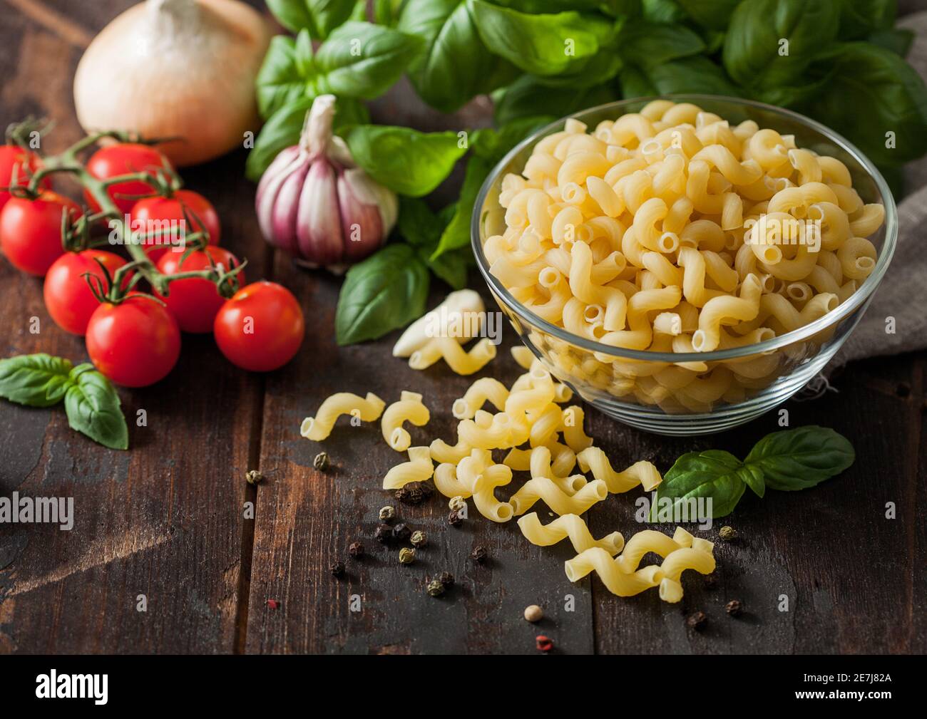 Raw maccheroni elbows pasta in glass bowl with oil and garlic, basil plant and tomatoes with pepper on wooden table. Top view Stock Photo