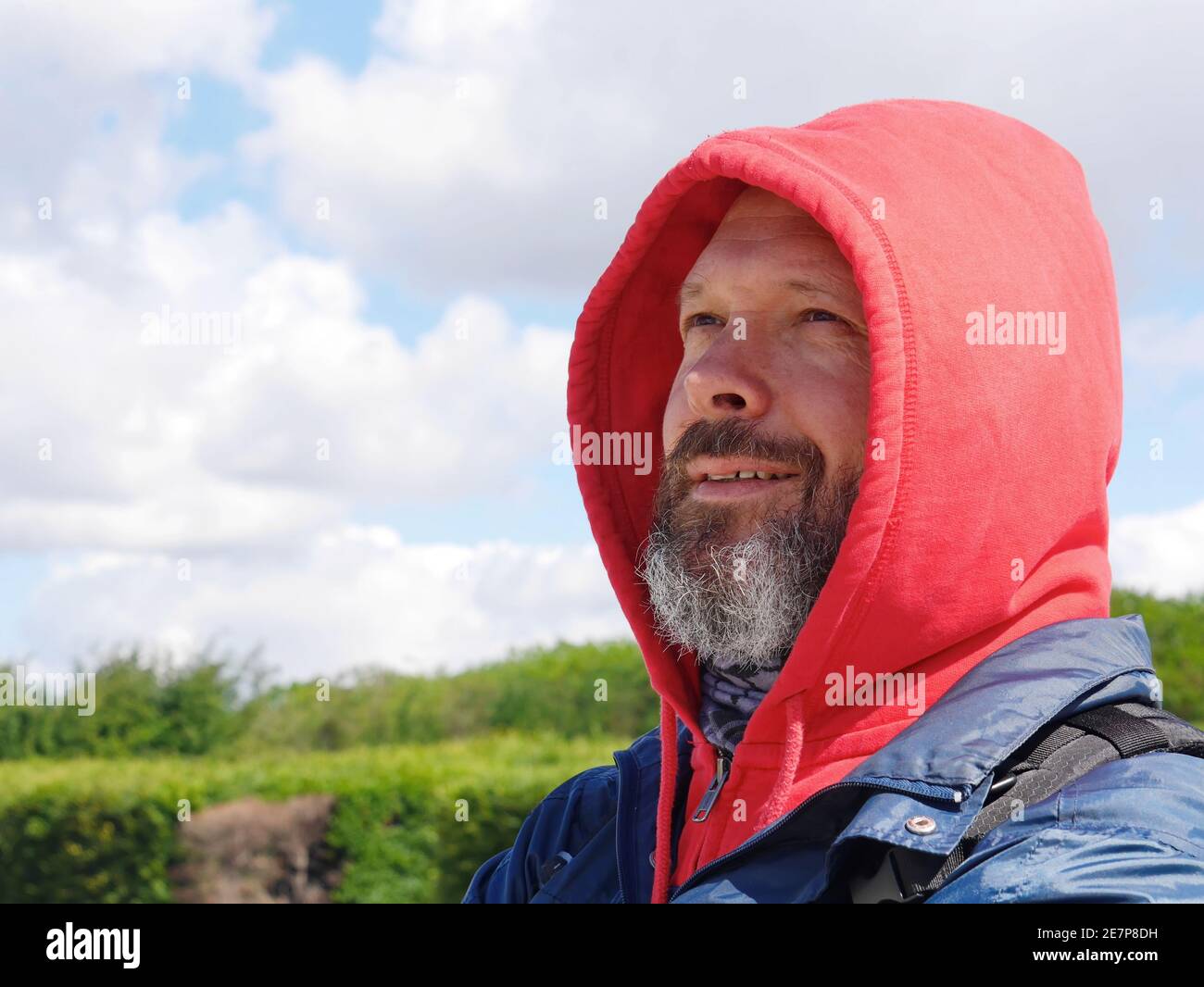 A bearded man in a red hood looks thoughtfully into the distance and smiles Stock Photo
