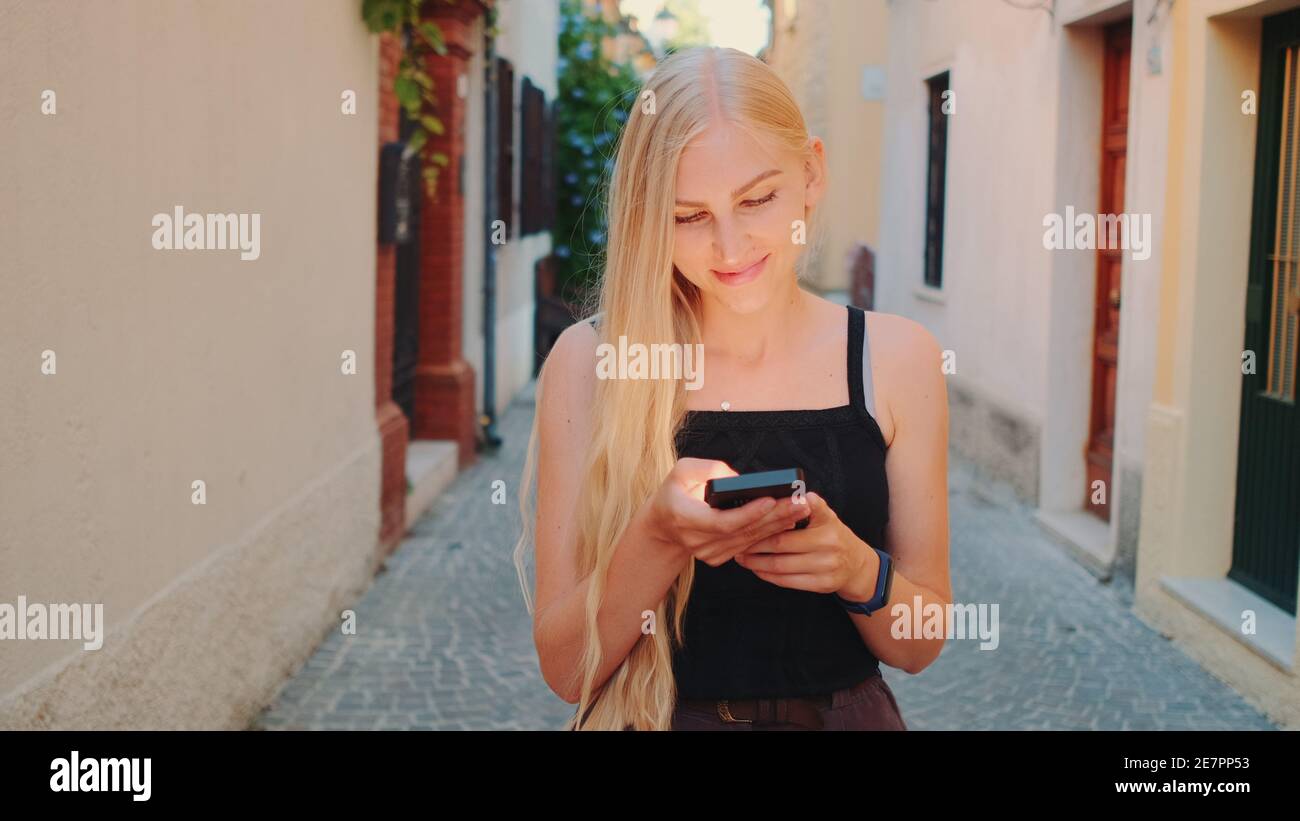 Pretty blonde girl chatting on smartphone while walking down the street of the old town. Smiling and happy woman. Stock Photo