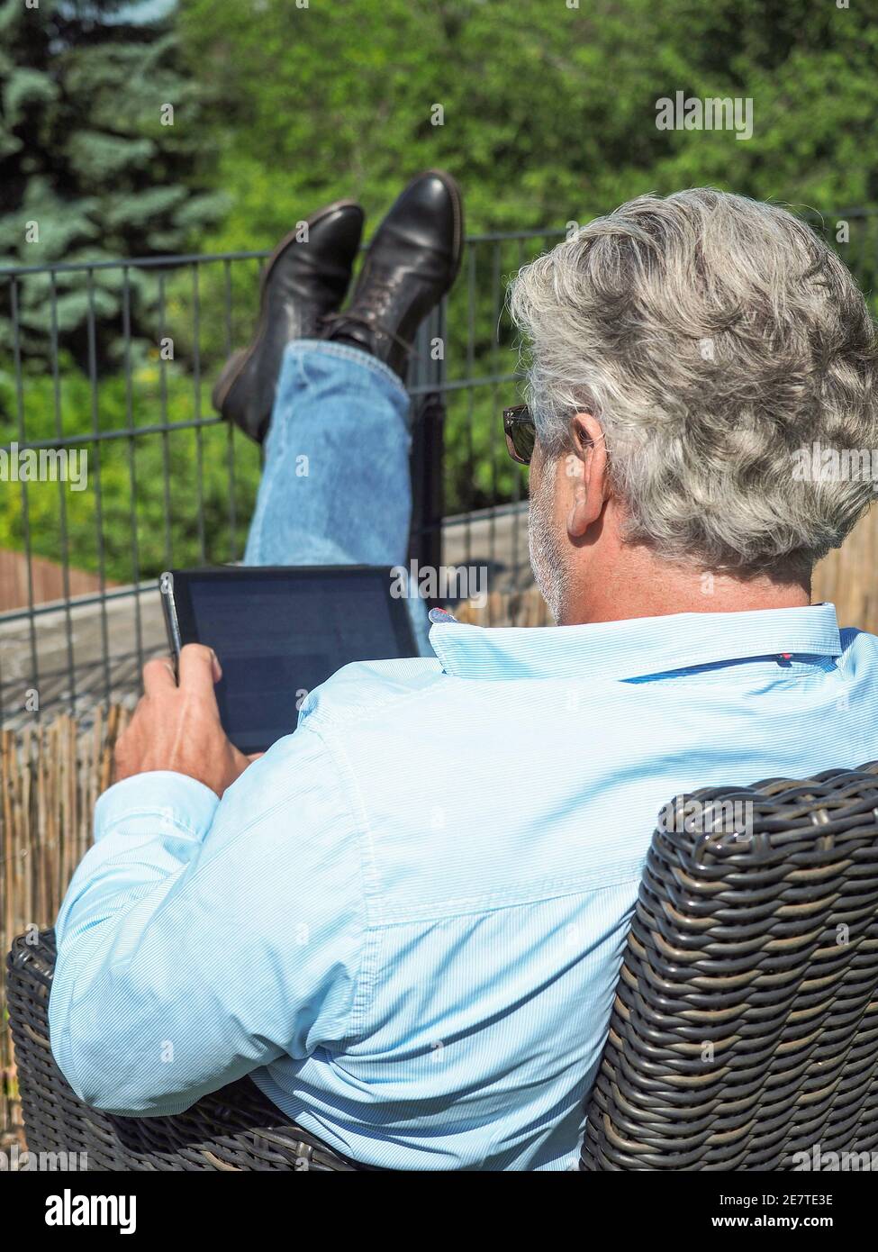 Senior man with grey hair sitting on a chair in the garden and looking to the tablet. From behind.  Stock Photo