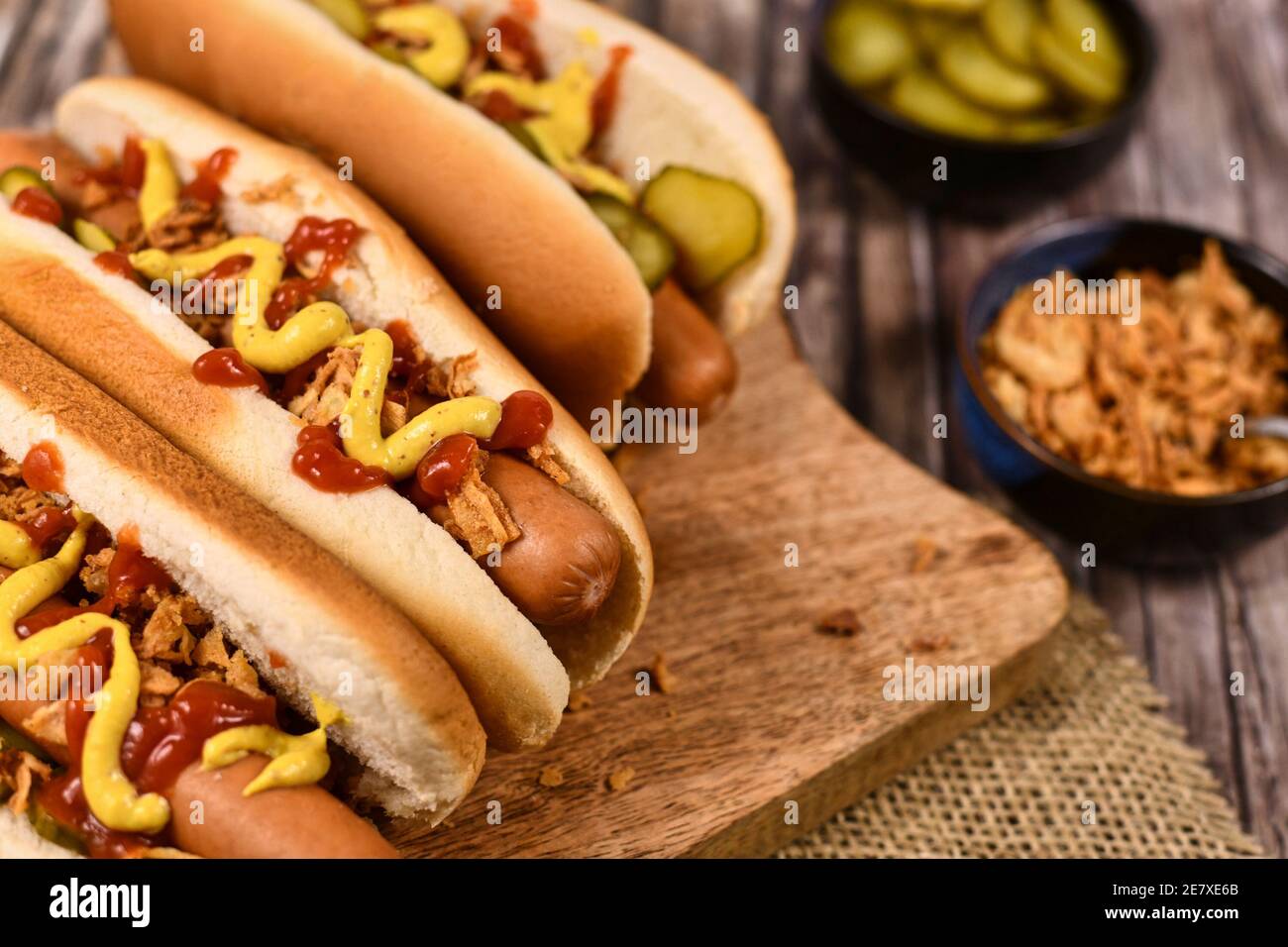 Homemade hotdogs with sausages and buns topped with preserved pickles, dried roast onions, mustard and ketchup next to bowls with ingredients Stock Photo