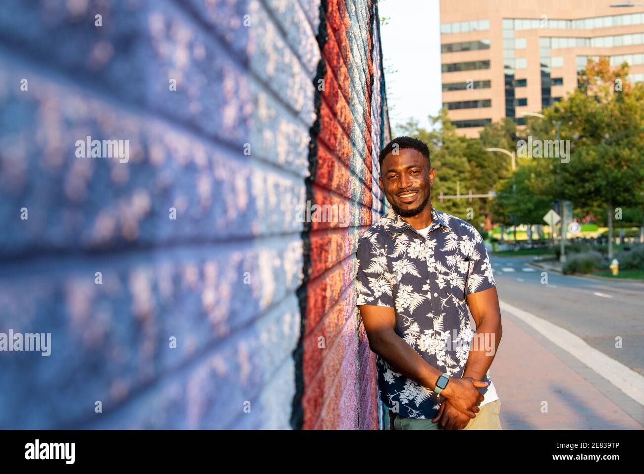 A Black man leans against a colorful street mural and smiles at the camera. Stock Photo