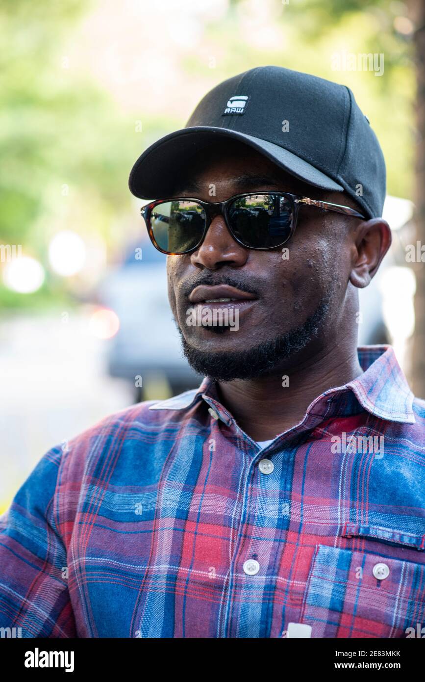 A Black man wears a basball cap and sunglasses in an urban setting. Stock Photo