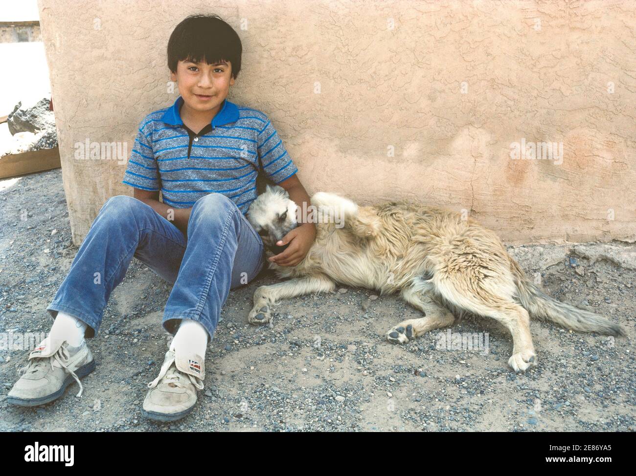 A young Native American boy and his pet dog rest in the shade at the side of his hilltop home in Acoma Pueblo, an American Indian community on a reservation west of Albuquerque, the capital of New Mexico in southwestern USA. Founded in the 12th Century, Acoma is one of the oldest continuously inhabited settlements in North America. The youngster is among the tribal members that live in rock and adobe dwellings atop a remote sandstone mesa. Tourists are welcome to join guided tours of his historic Sky City (Old Acoma) village. Stock Photo