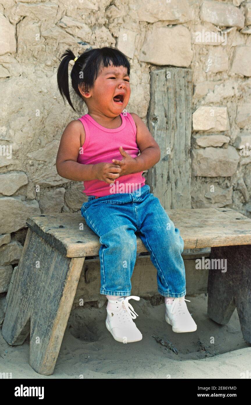 A young Native American girl cries as she sits on a bench in the shade at the Acoma Pueblo, an American Indian community on a reservation west of Albuquerque, the capital of New Mexico in southwestern USA. The youngster is among the tribal members that live in rock and adobe dwellings atop a remote sandstone mesa in the historic Sky City (Old Acoma) village. Founded in the 12th Century, Acoma is one of the oldest continuously inhabited settlements in North America. Stock Photo