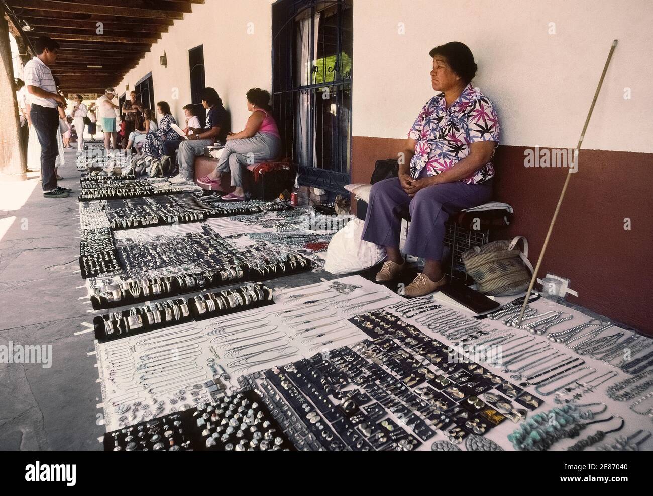 Native Americans and other local artisans sit in the shade behind their sidewalk displays of handmade jewelry awaiting tourists shopping for souvenirs in Old Town Plaza in Albuquerque, the capital of New Mexico, USA. This informal outdoor market on the east side of the plaza lines the covered patio in front of historic La Placita Dining Rooms that has been serving traditional New Mexican food since the 1930s. Stock Photo