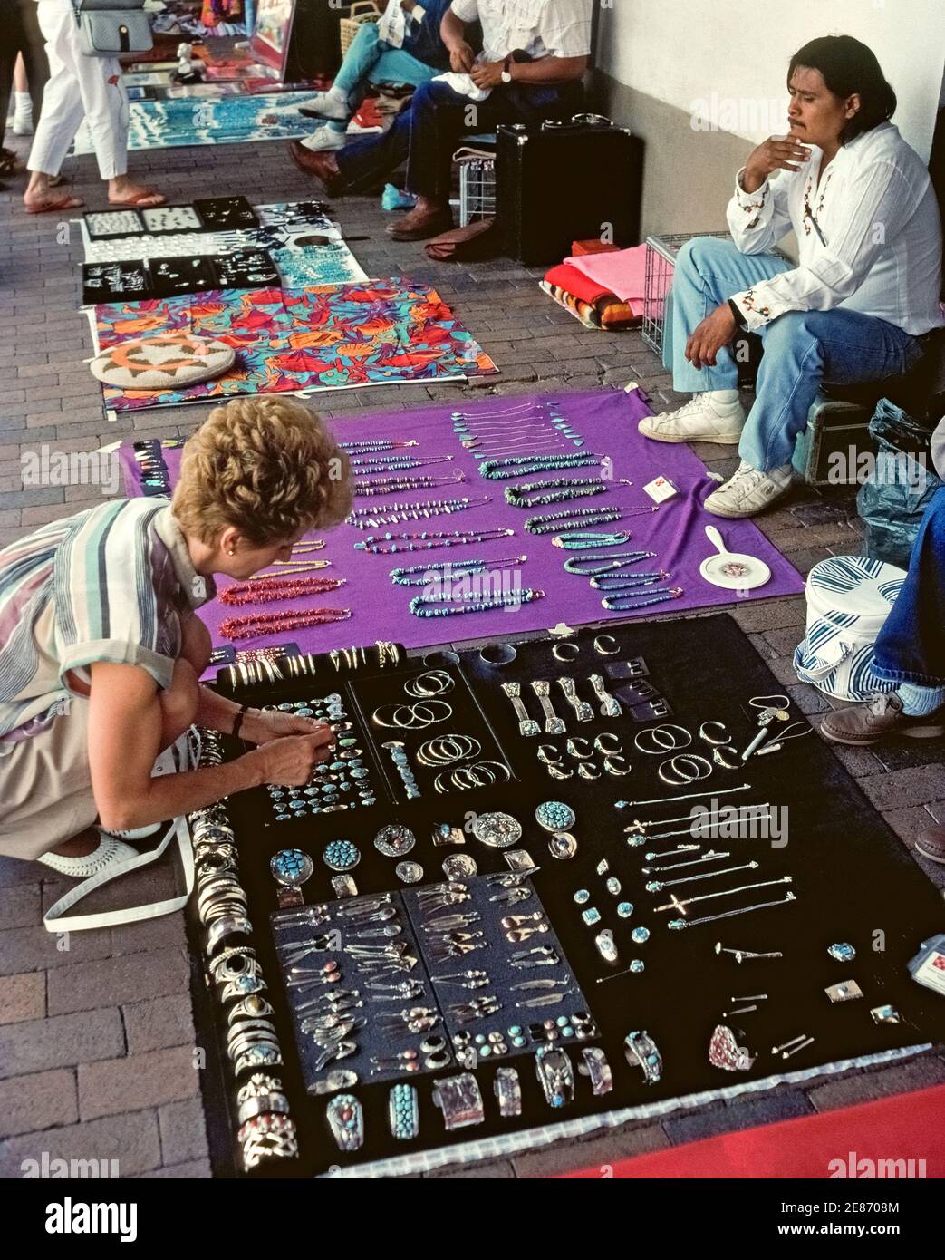 A female tourist stops to buy some Indian sterling silver jewelry displayed on the sidewalk at an outdoor market of Native American handicrafts at the downtown plaza in Santa Fe, New Mexico, USA. For more than a half century, American Indian artisans have been selling jewelry, beadwork, weavings, pottery and other handmade arts and crafts while sitting under the Palace of the Governor portal on the north side of plaza. The Palace is home to the New Mexico History Museum and a National Historic Landmark. Stock Photo