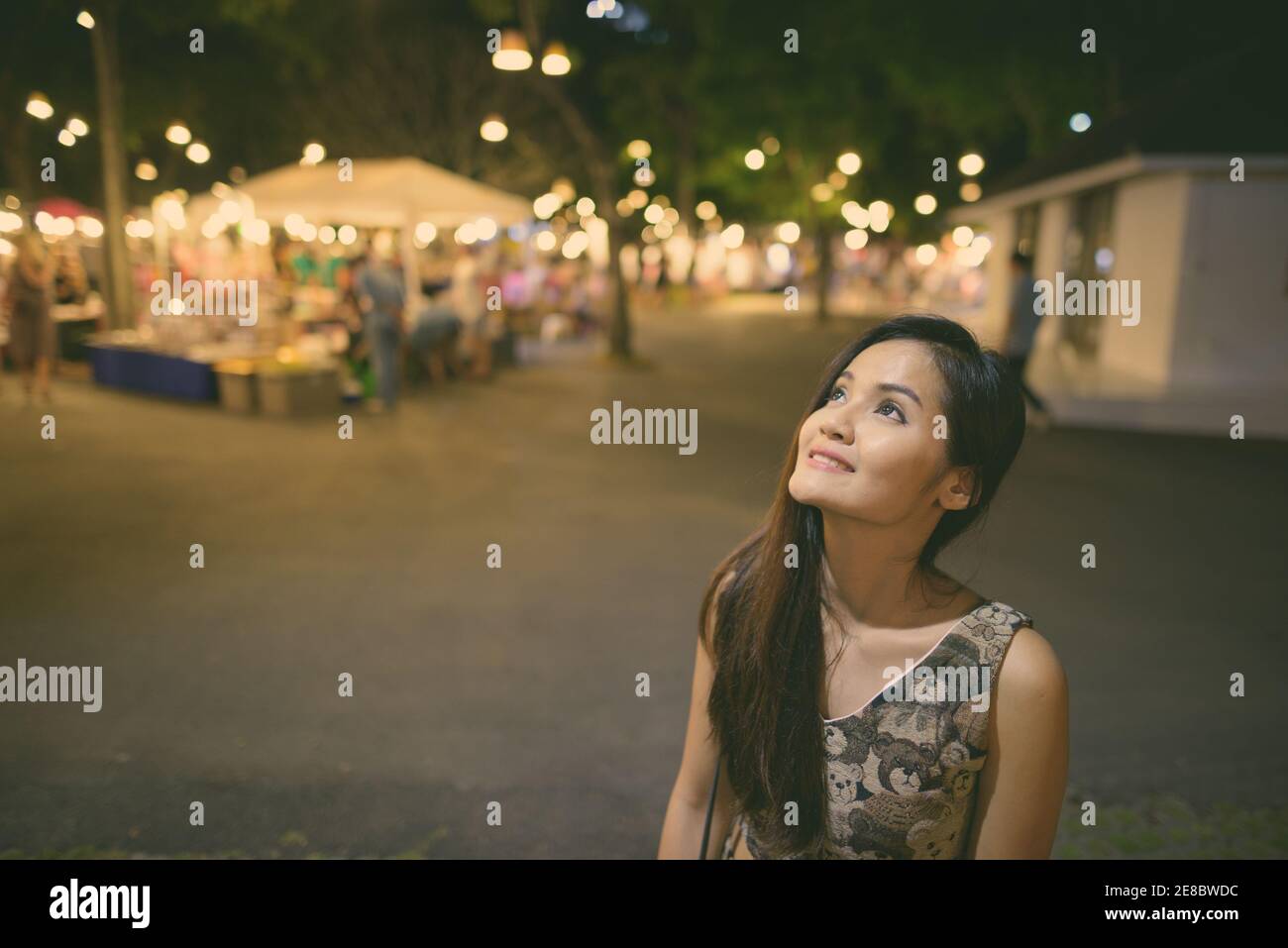 Young happy beautiful Asian woman smiling and thinking against the night market in Hua Hin Thailand with people buying and selling various goods Stock Photo