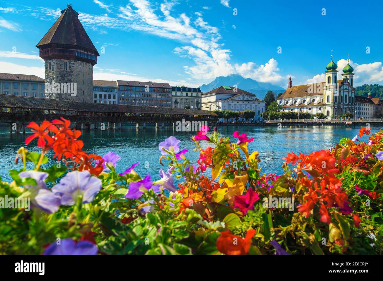 Amazing touristic and travel destination in Lucerne. Cute Jesuitenkirche church with Reuss river. Waterfront view with flowered wooden Chapel bridge, Stock Photo