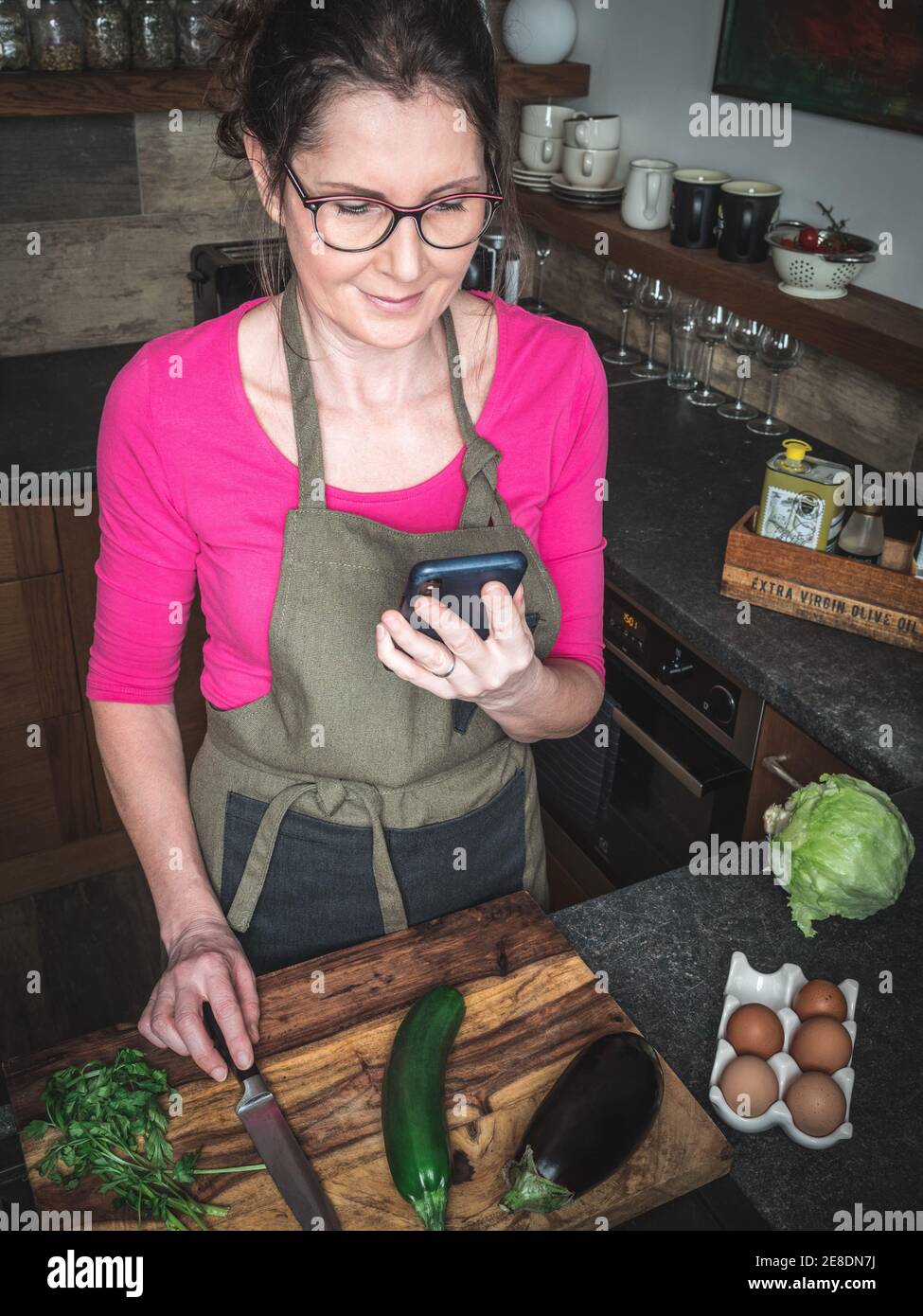 Caucasian middle aged woman in the kitchen follows recipe on smartphone. Healthy food concept. Stock Photo
