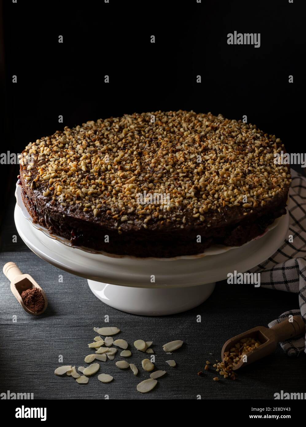 A cocoa cake filled with fruit jam and covered with chopped hazelnuts on a white stand and black background Stock Photo