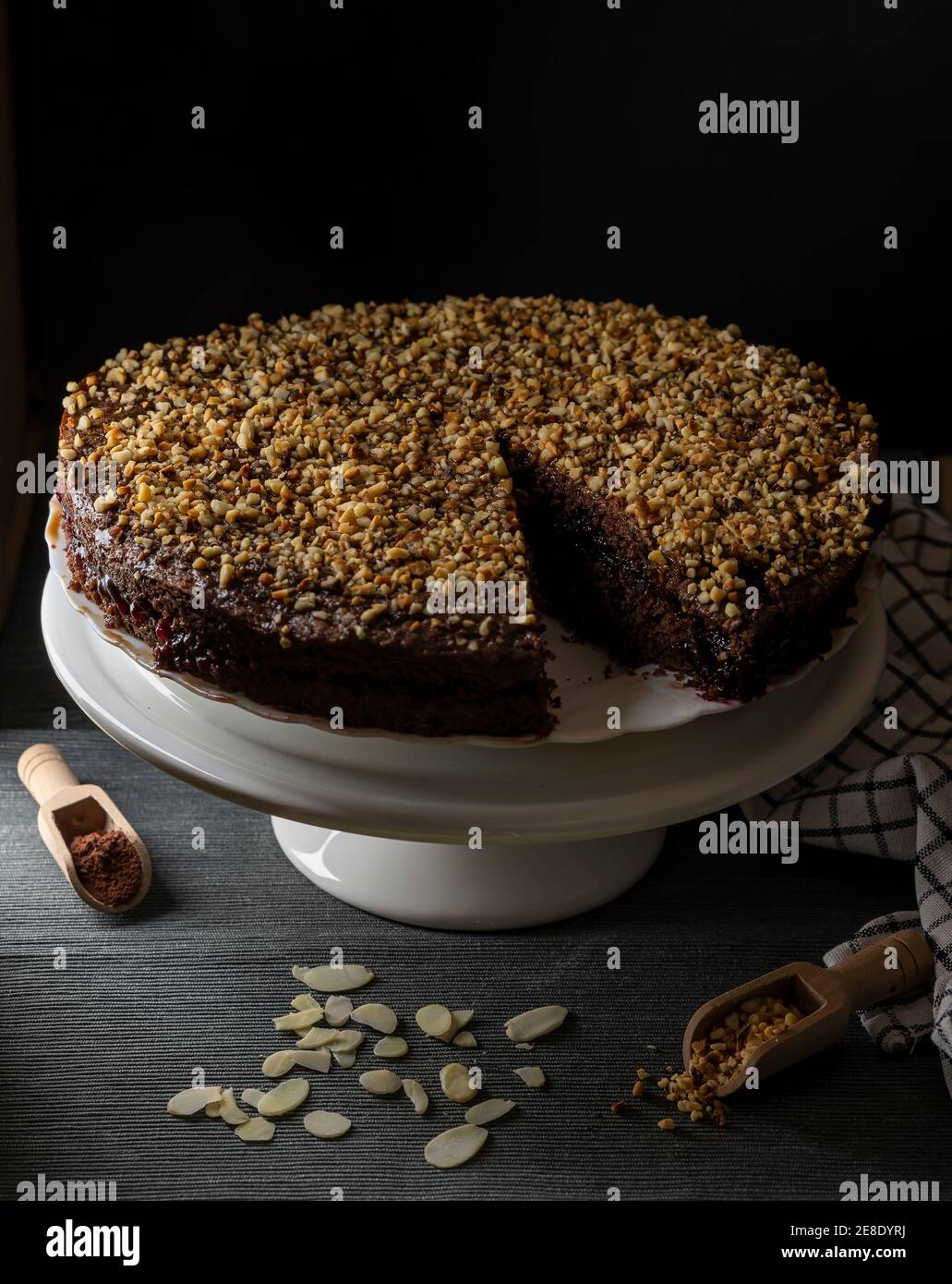 A cocoa cake filled with fruit jam and covered with chopped hazelnuts on a white stand and black background Stock Photo