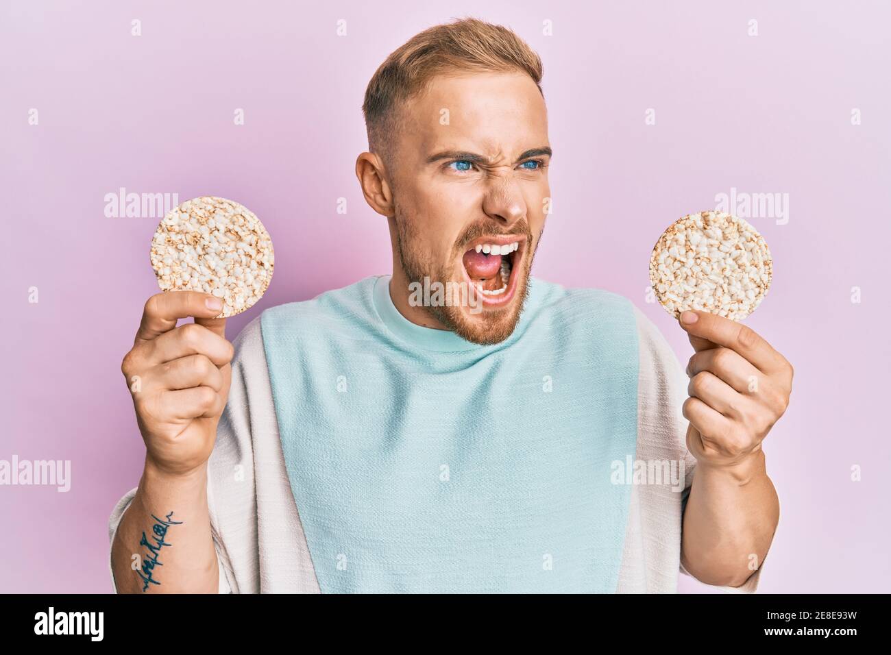 Young caucasian man eating healthy rice crackers angry and mad
