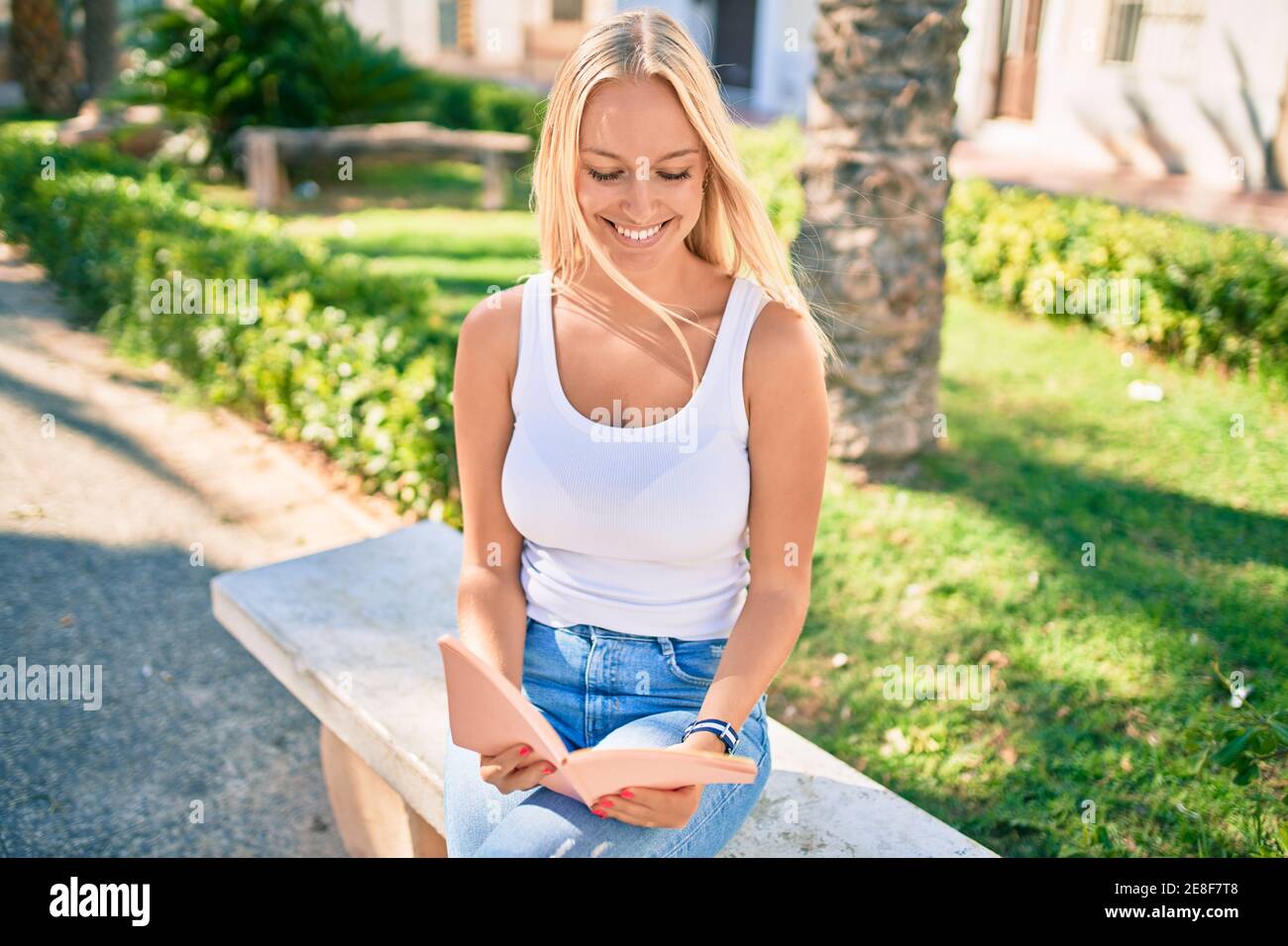 Young blonde girl smiling happy reading book sitting on the bench at ...