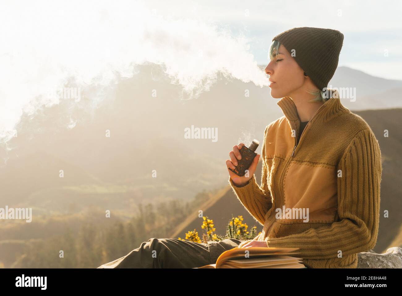Side view of peaceful female traveler sitting on rock while smoking vape and exhaling cloud of smoke during vacation in mountains Stock Photo