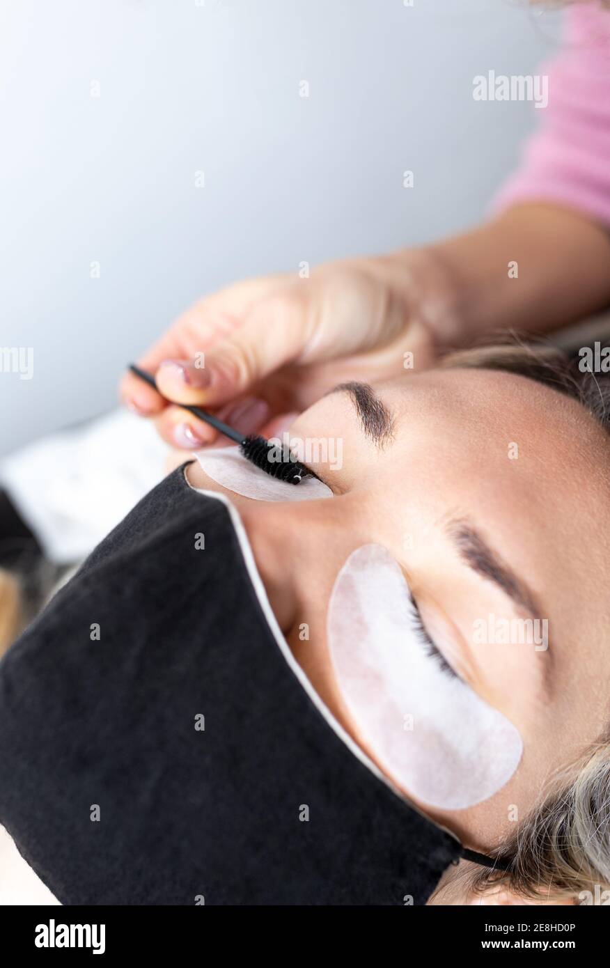 Professional beautician applying artificial eyelashes on young female client wearing protective face mask in modern beauty studio Stock Photo