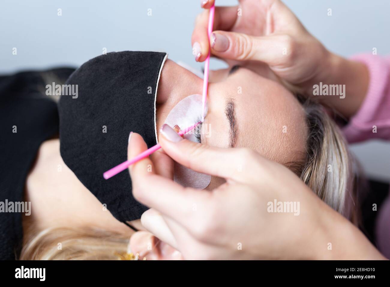 Professional beautician applying artificial eyelashes on young female client wearing protective face mask in modern beauty studio Stock Photo