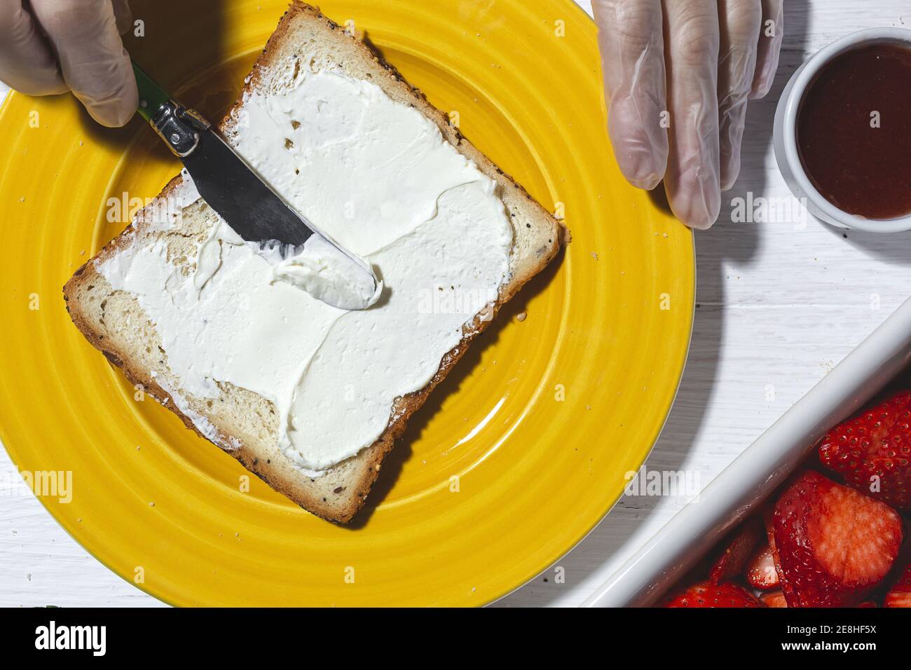 Top view crop anonymous female cook in latex gloves spreading yummy cream cheese on bread slice placed on yellow plate near cut strawberries Stock Photo