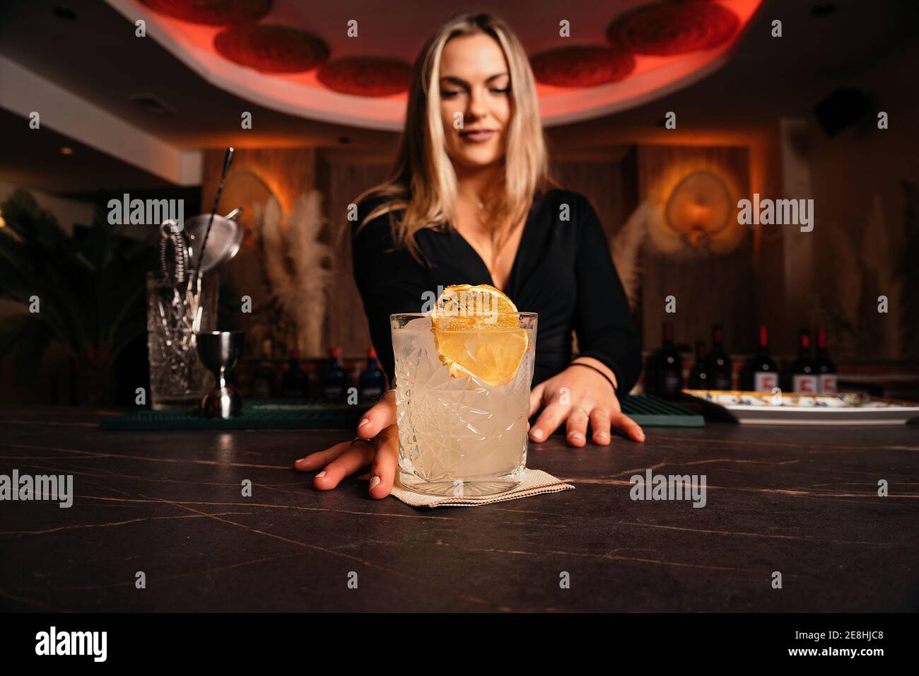 Content young female bartender with long blond hair serving cold alcohol cocktail decorated with lemon slice in stylish restaurant Stock Photo