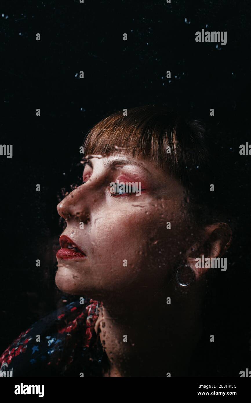 Unemotional female with ear piercing standing behind translucent glass with water droplets looking away Stock Photo