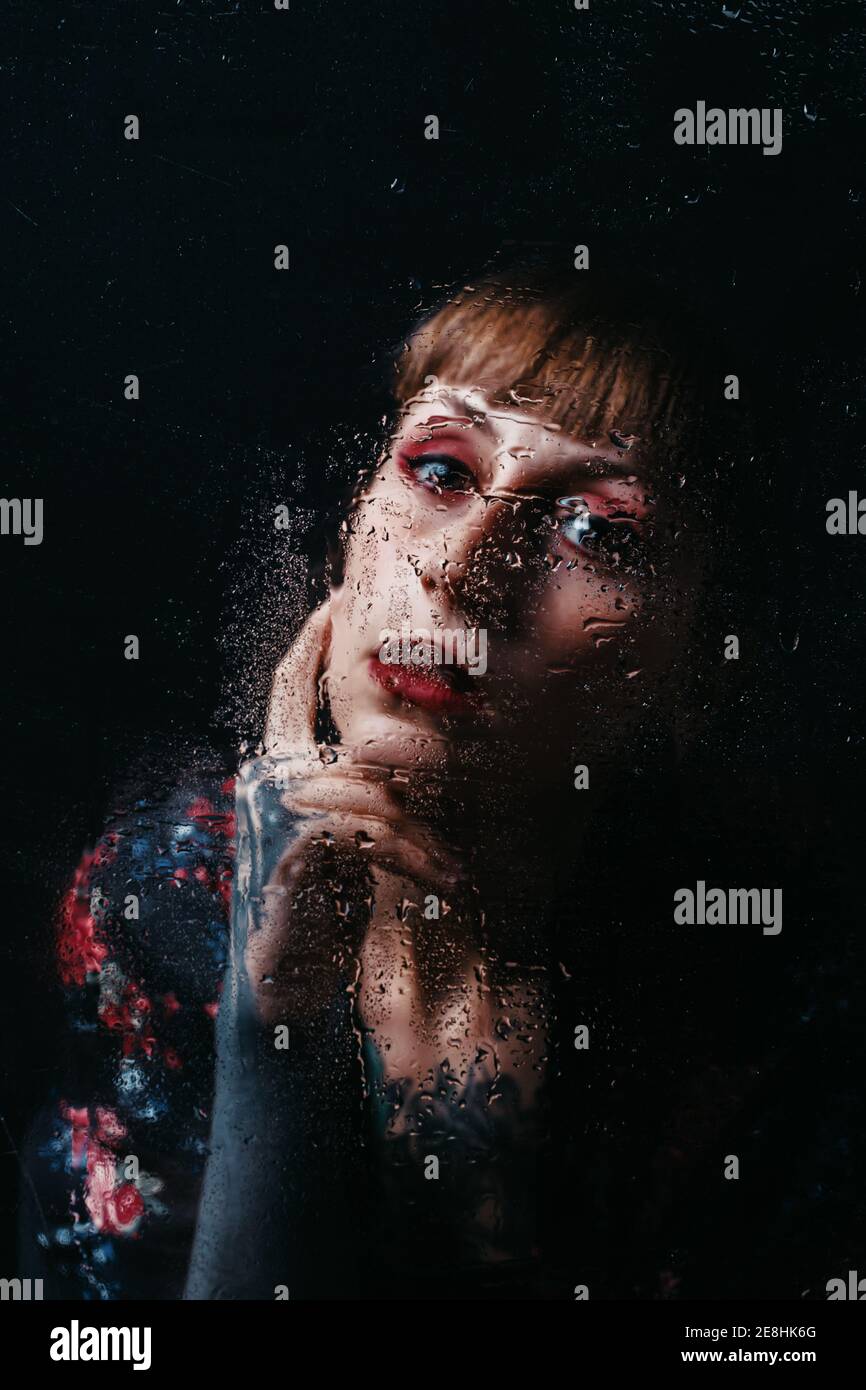 Unemotional female with arm tattoo touching neck standing behind translucent glass with water droplets looking at camera Stock Photo