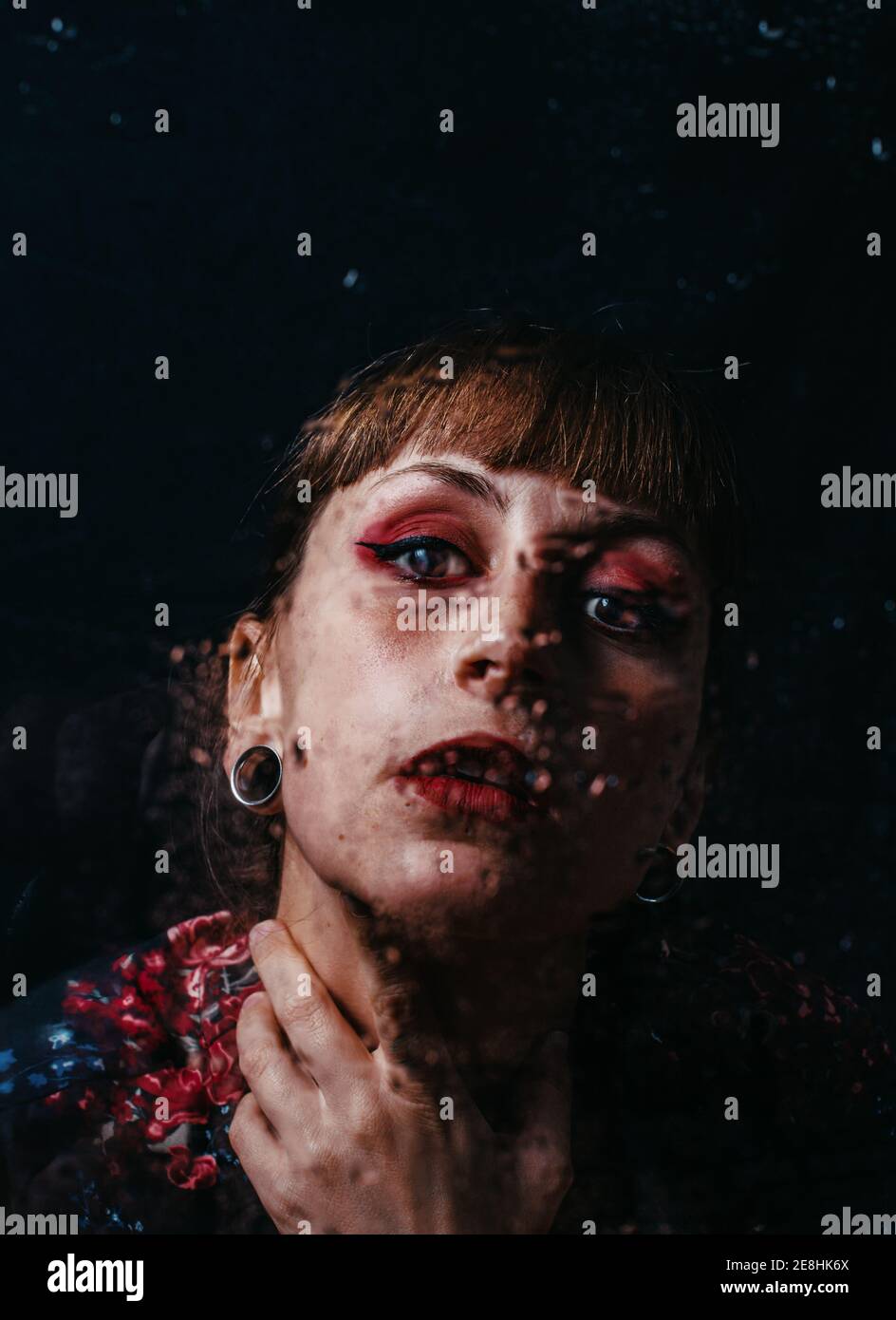 Unemotional female with arm tattoo touching neck standing behind translucent glass with water droplets Stock Photo