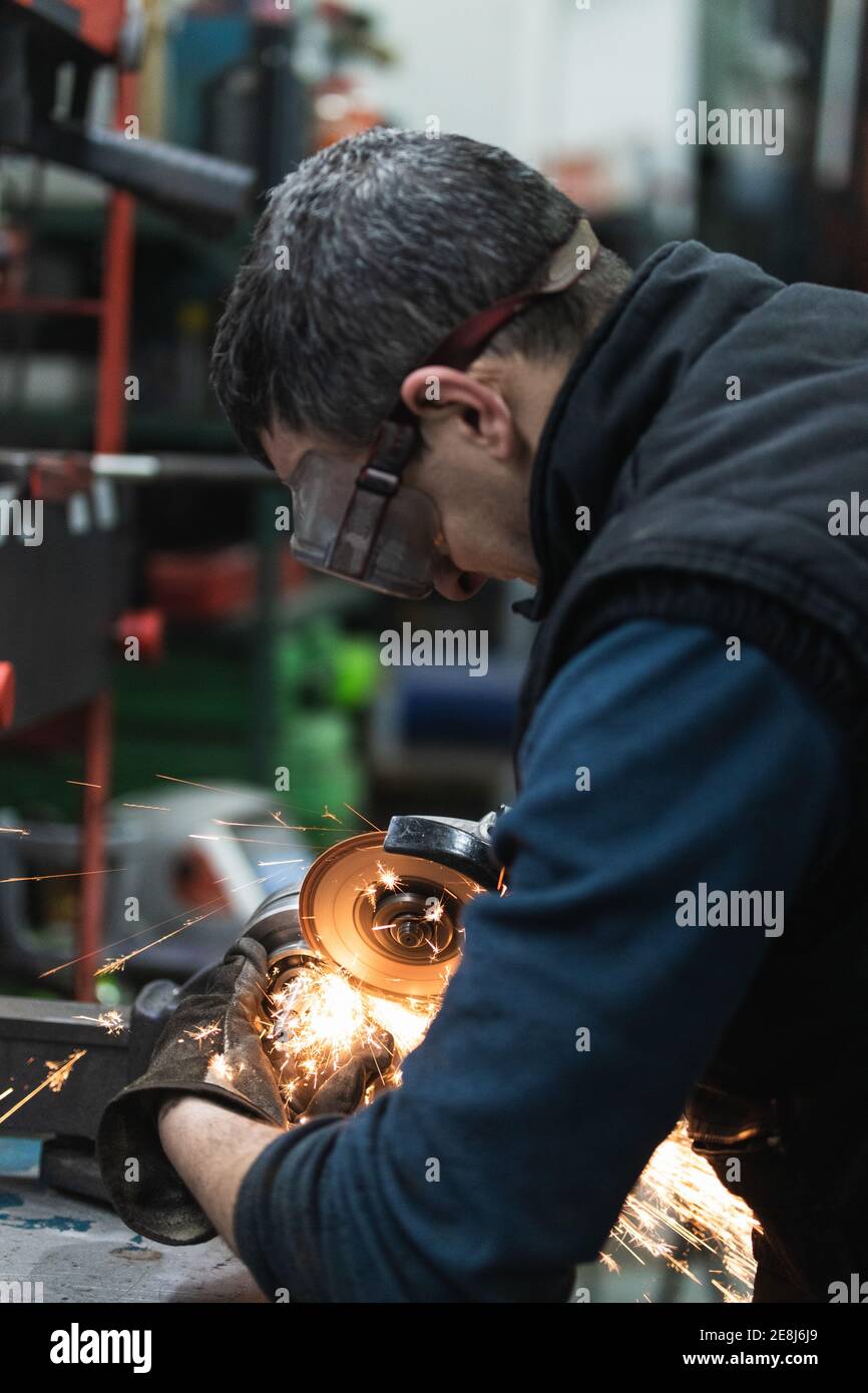 Side view of crop adult male mechanic in casual clothes and protective goggles and gloves cutting metal detail with angular grinding tool during work Stock Photo