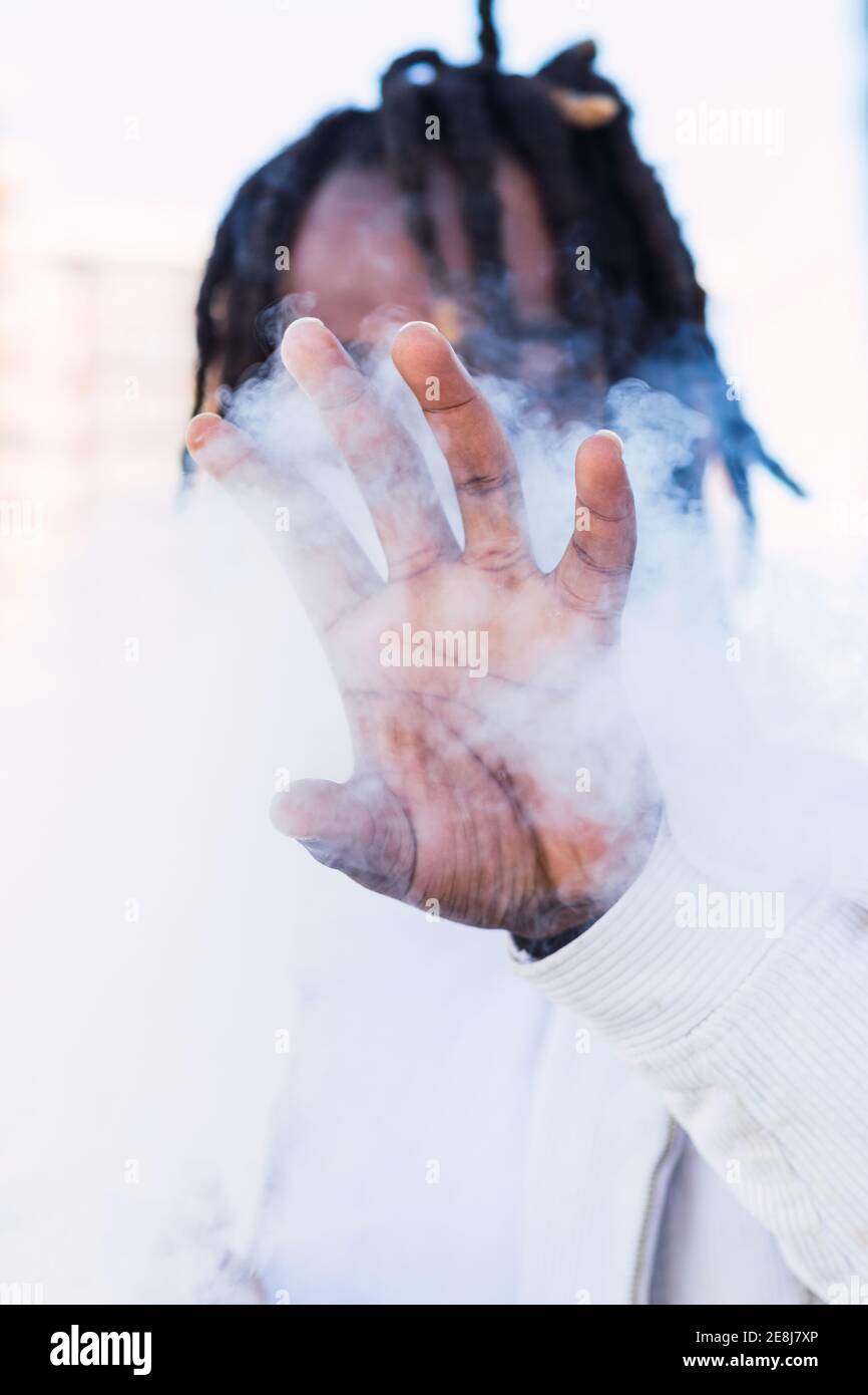 Unrecognizable African American male with dreadlocks covering face with hand while standing in street in cloud of fume and smoking electronic cigarett Stock Photo