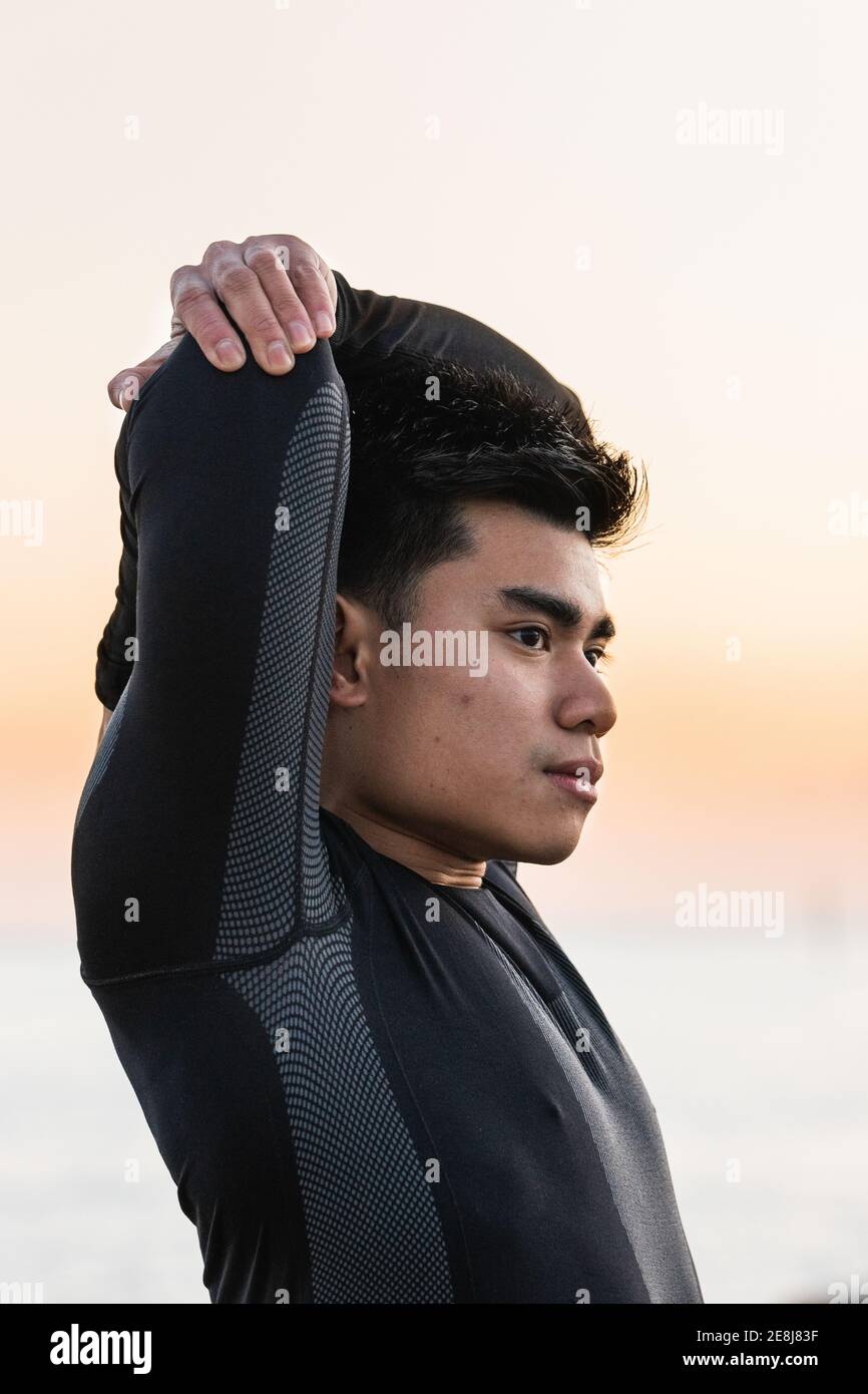 Side view of young Asian male in sportswear stretching arms and shoulders while standing against blurred sea and sunset sky during outdoor fitness wor Stock Photo