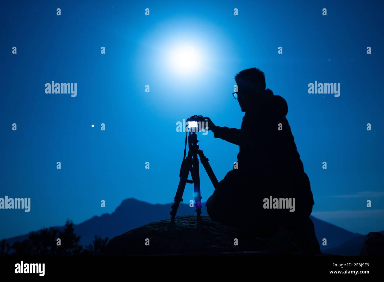 Side view of unrecognizable male traveler silhouette with photo camera on tripod against ridge under blue sky with shiny sun at night Stock Photo