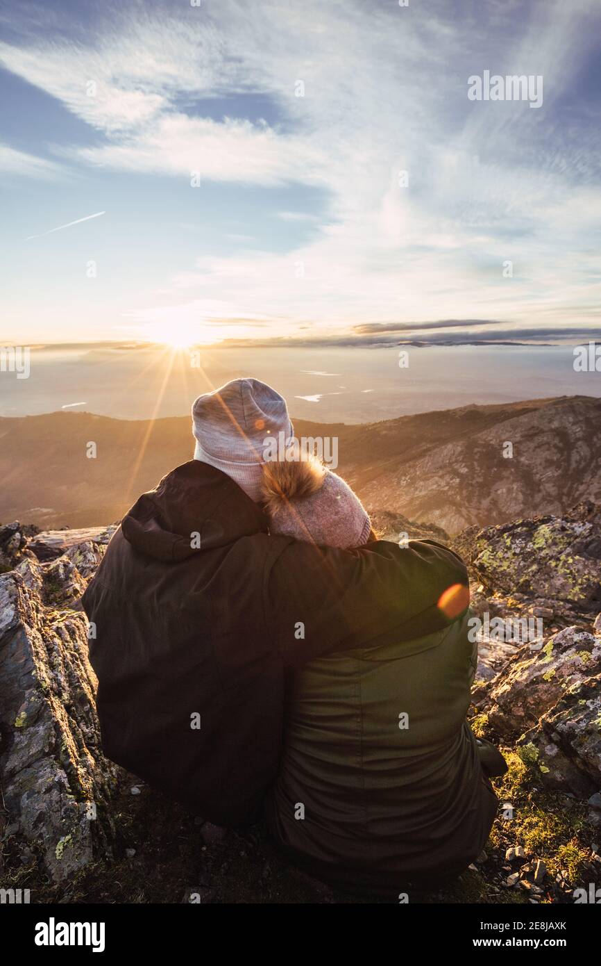 Back view of unrecognizable man embracing girlfriend in outerwear while admiring dawn from mountain under cloudy sky in sunshine Stock Photo