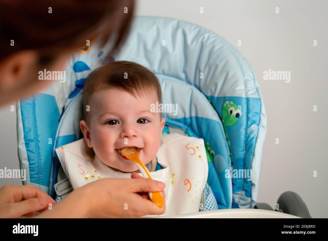 Adorable happy kid wearing bib sitting in stroller and getting fed with sweet baby food by