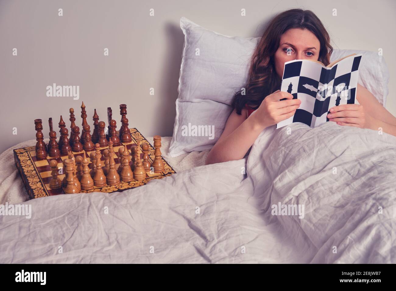 A woman reads a chess book at night on the bed next to the chessboard. Stock Photo