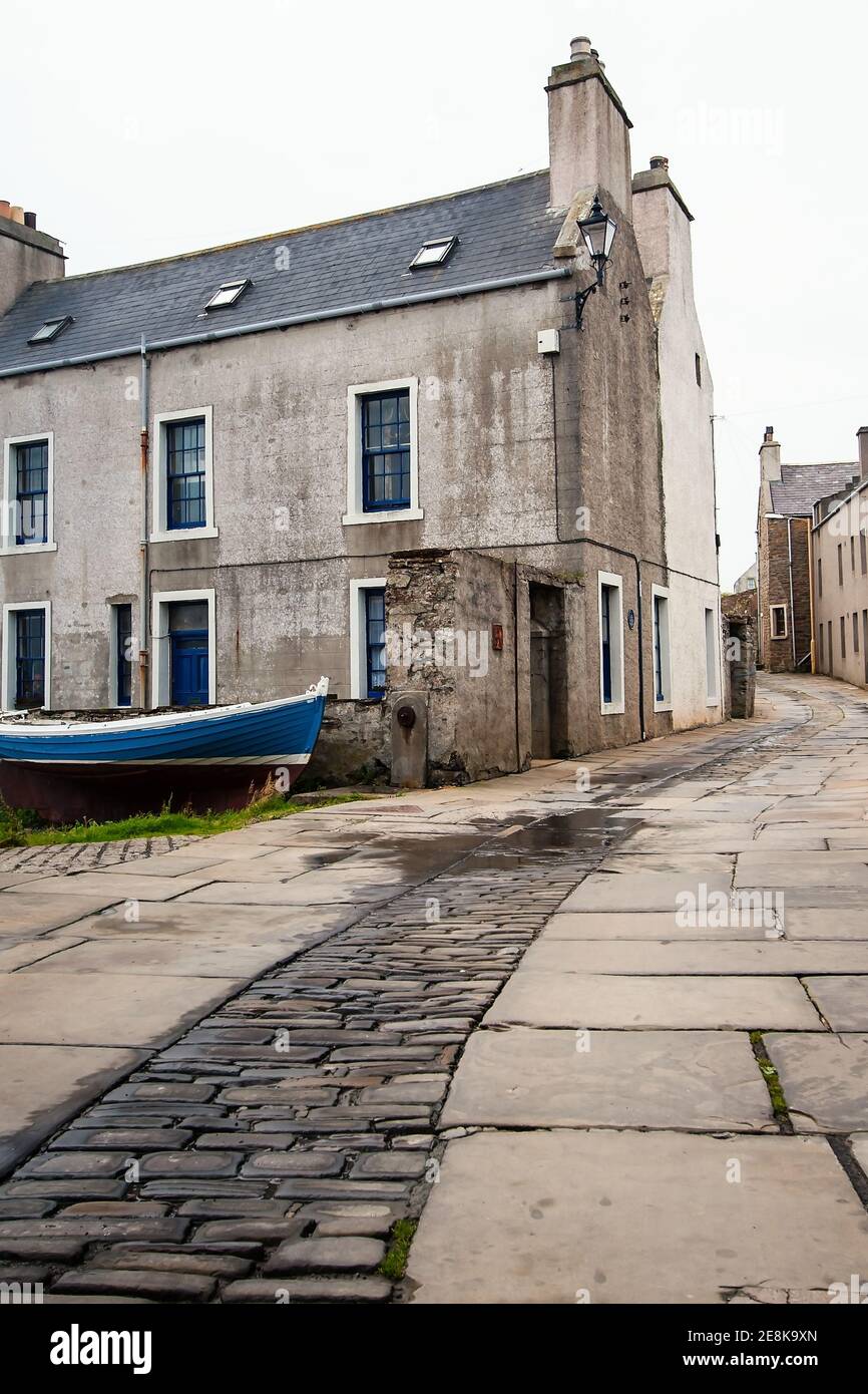 Narrow pebble and stone slabs street in northern Scotland town on Orkney islands with blue boat in foreground and big stone building Stock Photo