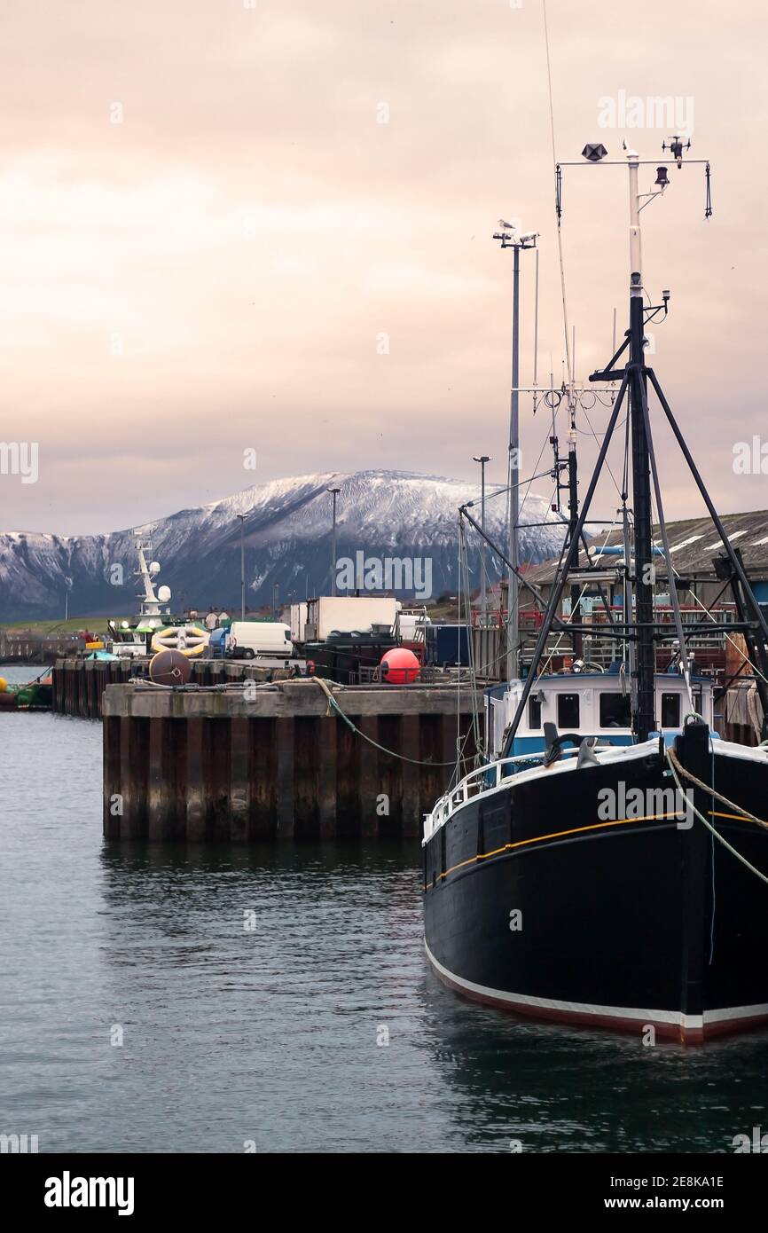 Fishing harbour on Orkney in winter scene with boat in foreground and snow on hill in background in Stromness Stock Photo
