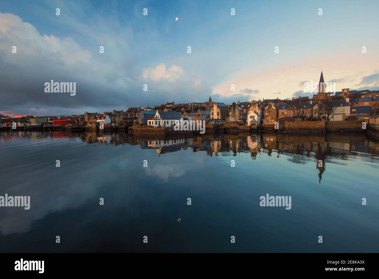 Wide angle view of fishing town Stromness on Orkney islands with moon reflecting in water and distandt stone buildings at sunrise Stock Photo