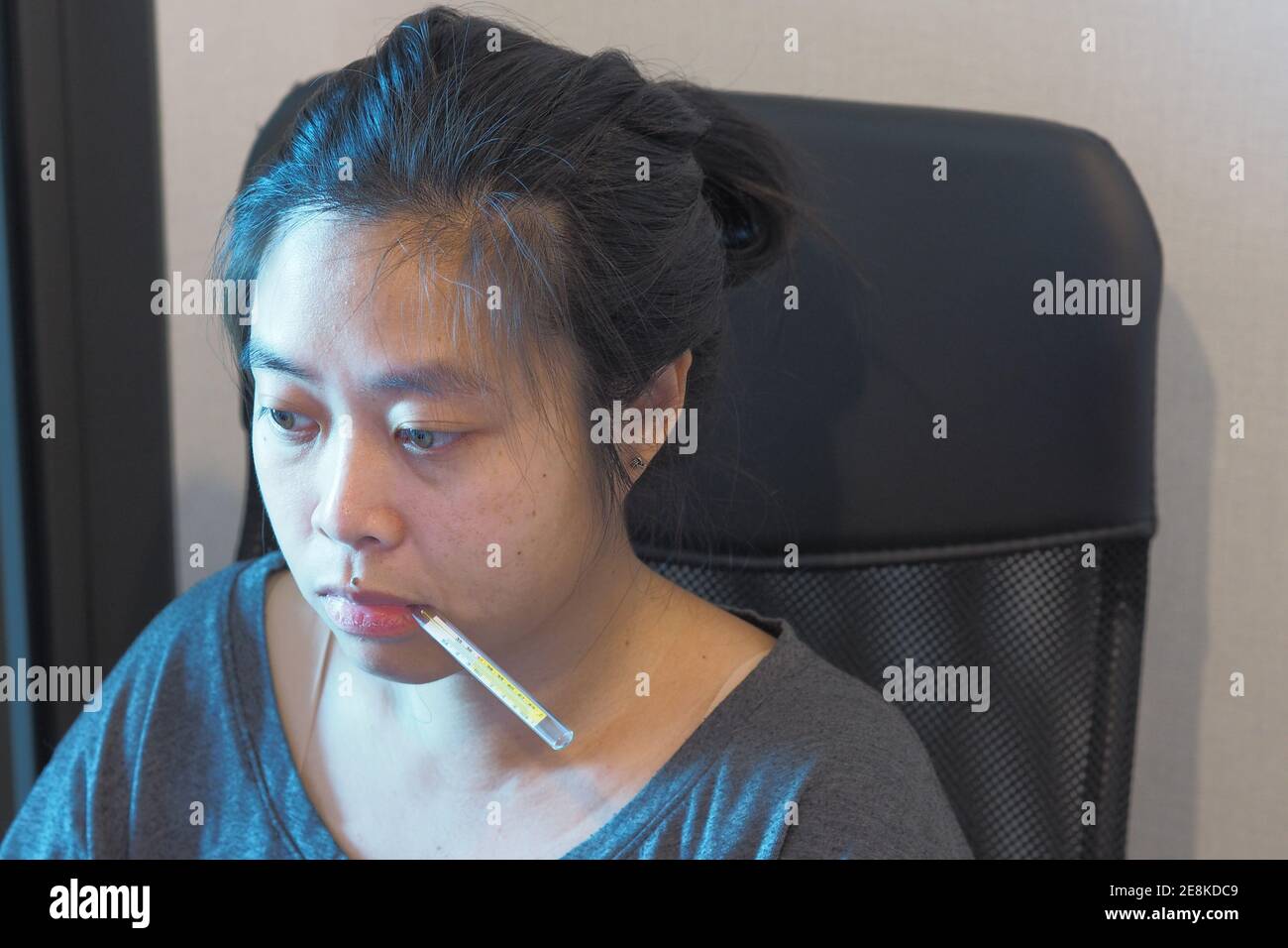 Asian Woman with thermometer in her mouth on office chair - Work from home concept. Stock Photo