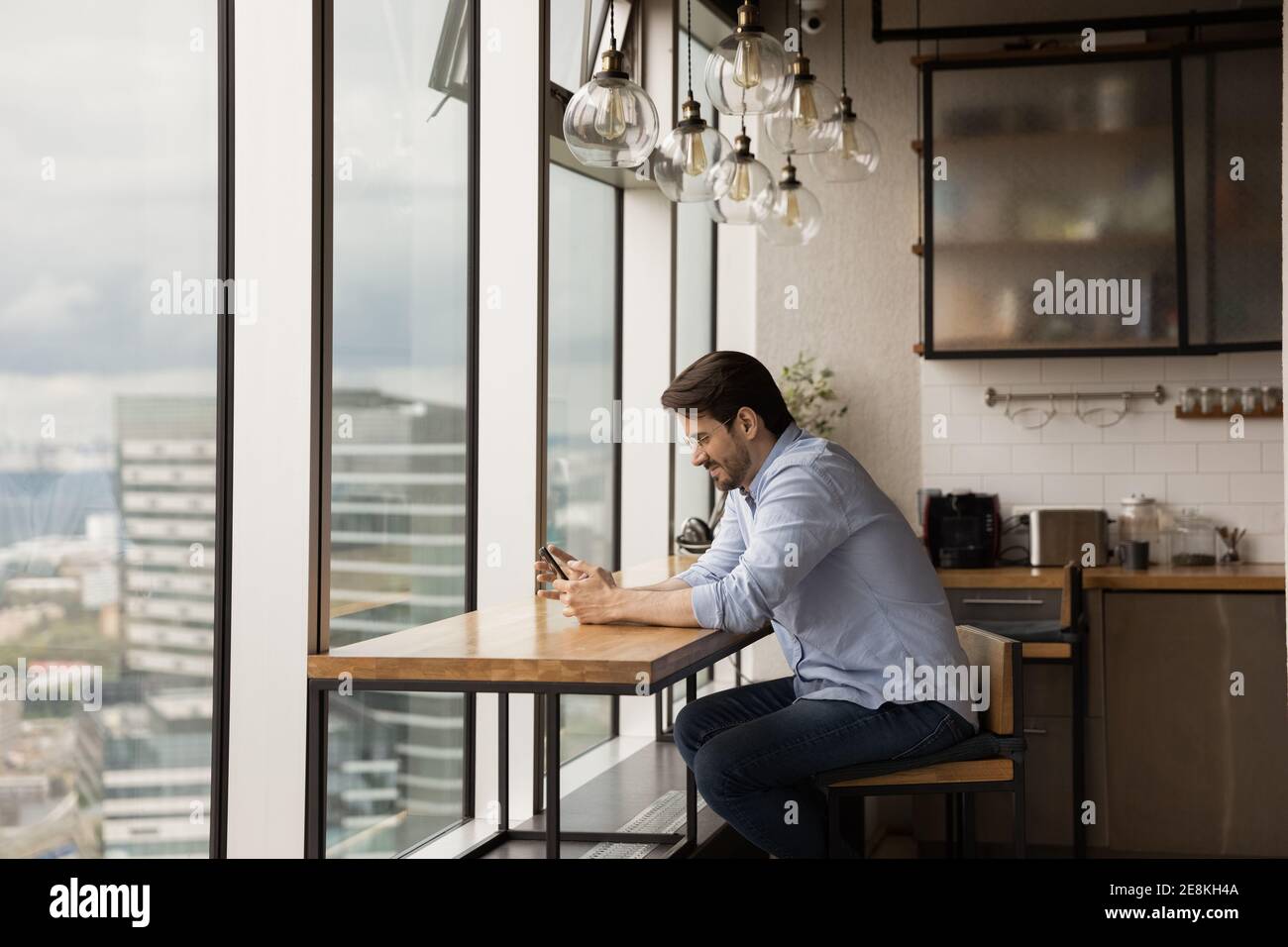 Smiling young man relaxing near big panoramic window using smartphone Stock Photo
