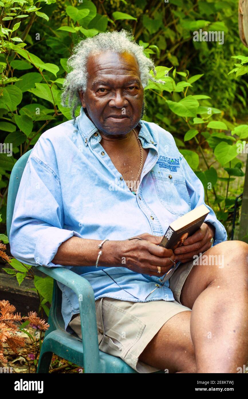 Canadian-Barbadian writer Austin Chestfield (1934-2016) holding his novel More and sitting in his backyard in summer Stock Photo