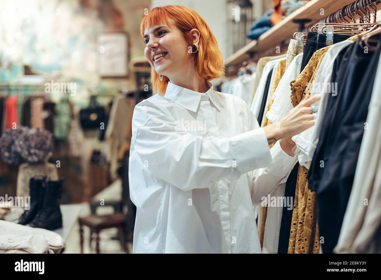 Attractive woman choosing clothes in shop. Female customer looking at fashionable clothes in a store and smiling. Stock Photo