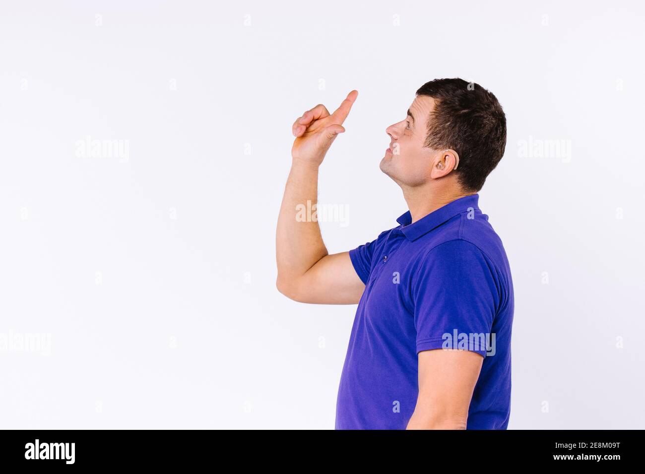 Sign language. Caaucasian man wearing casual clothes looks up and points up with index finger against white background with side ad space. Stock Photo