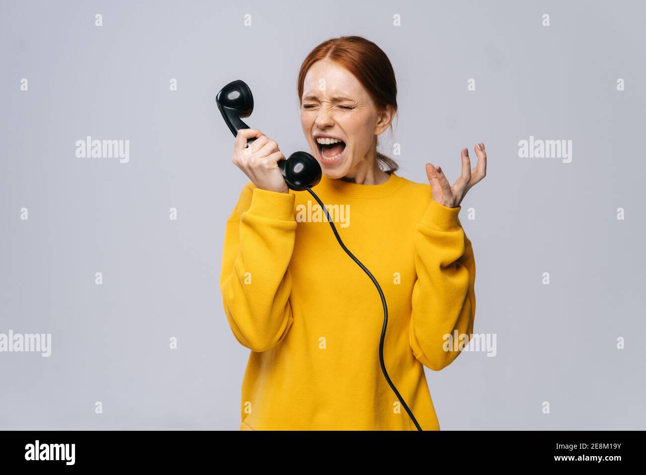Crazy angry young woman in stylish yellow sweater talking on retro phone and screaming in handset. Stock Photo