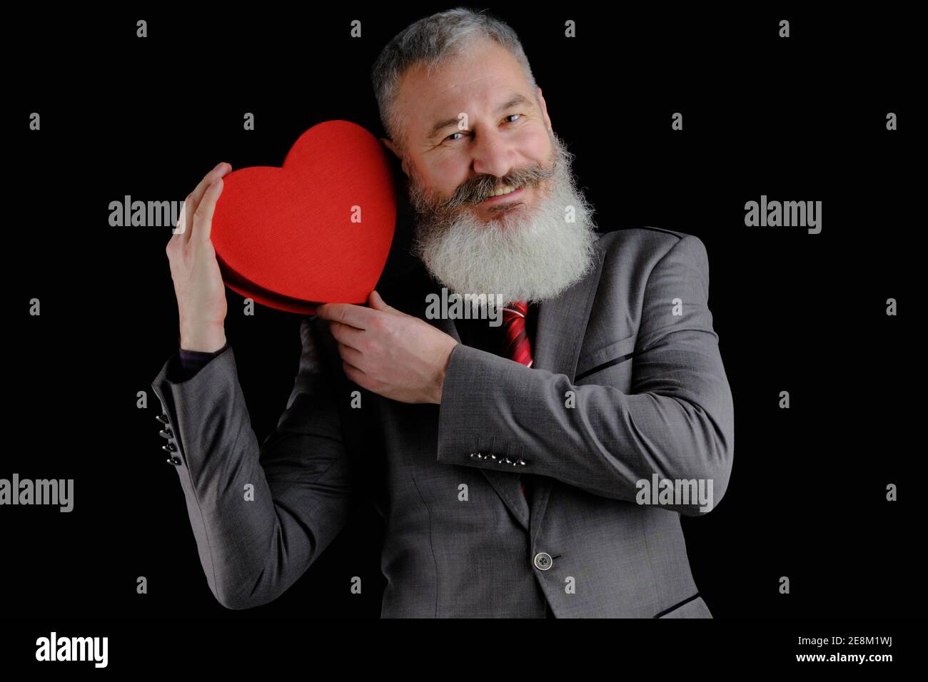 Mature bearded man wear gray suit holds red heart shaped gift box, isolated black background Stock Photo