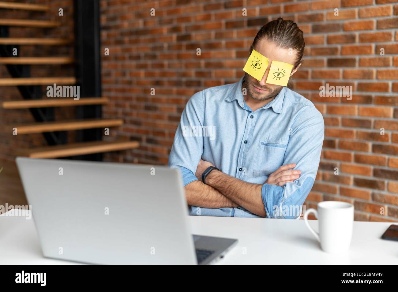 A young tired bearded office worker in a casual shirt sitting at the desk at his workplace, put stickers on the eyes to hide that he is napping or Stock Photo