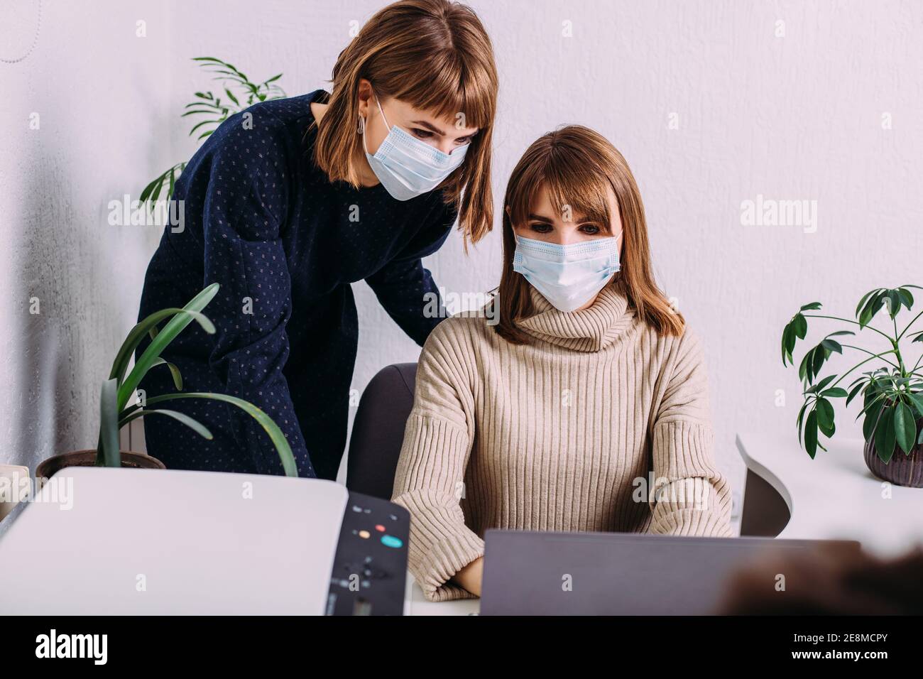 Beautiful twins sisters women with a medical protective face mask working together in a light office surrounded by plants. Stock Photo