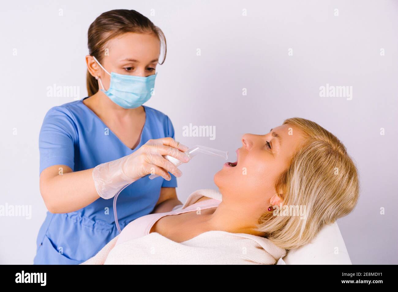 Young doctor in a medical mask and latex gloves treats the throat of an older woman with nebulizer on a white background. Fighting virus colds. Stock Photo