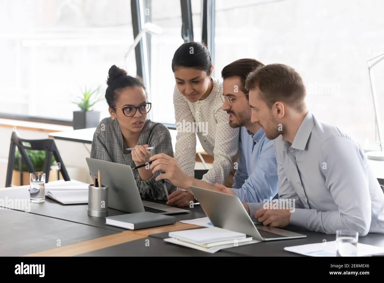 Mixed raced millennial employees talking at shared desk with laptops Stock Photo
