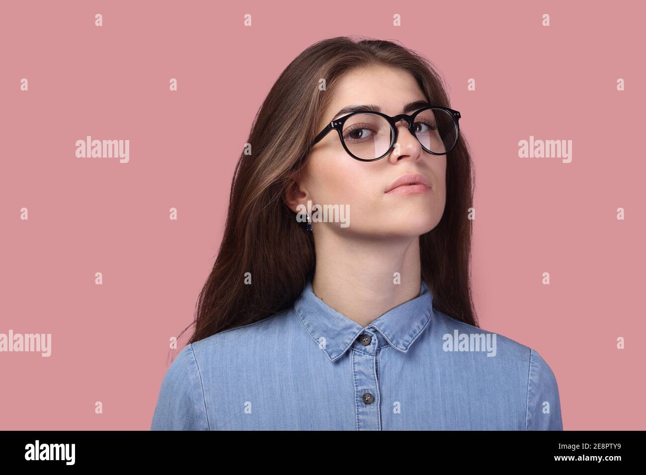 Attractive pensive woman with long hair in glasses and looking at camera, has pleasant appearance, isolated over pink background. Stock Photo