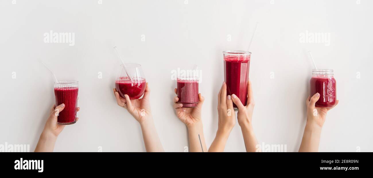 Human hands holding glasses and jar with purple beet pomegranate fresh smoothie with glass straws over white wall background, wide composition. Detox, dieting, weight loss, healthy lifestyle concept Stock Photo
