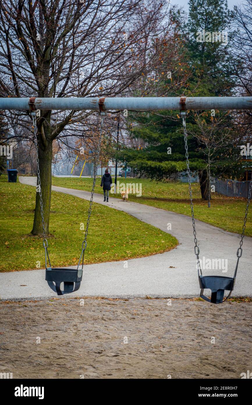 Woman alone walking her dog in a deserted children playground with empty swings during the pandemic Stock Photo